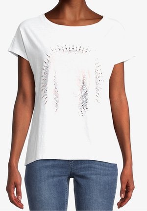 Vrouw draagt een wit shirt met korte mouwen en een vaag geometrisch zonnestraalpatroon, gecombineerd met een blauwe spijkerbroek.