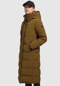 Long, padded coat in olive green with a high collar, black zipper, and side pockets. Features a drawstring hood and logo patch on arm.