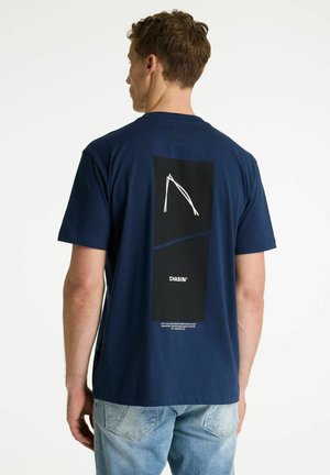 Mężczyzna w granatowej koszulce z czarnym nadrukiem, białym abstrakcyjnym wzorem i napisem "CHASIN'" na plecach, w połączeniu z jasnoniebieskimi dżinsami.