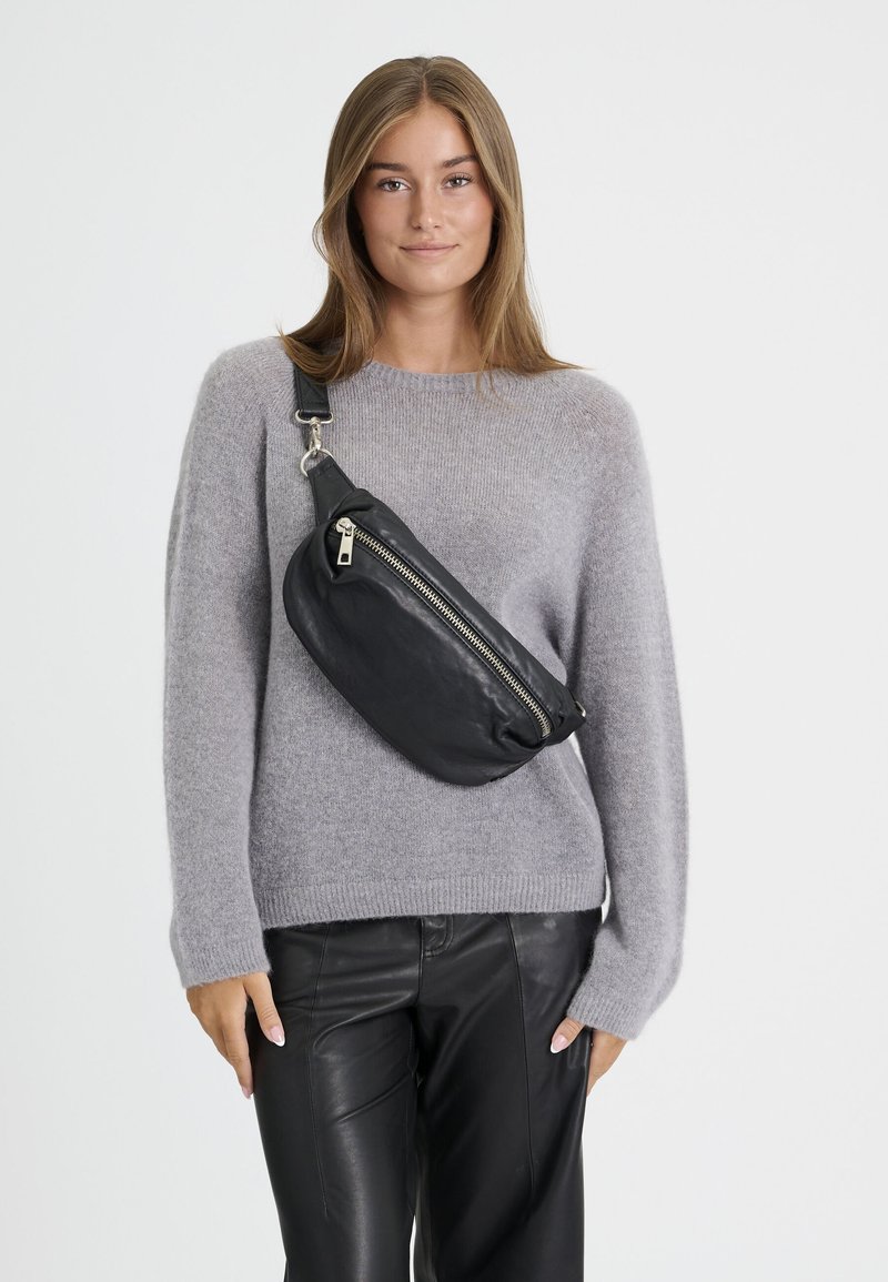 Jeune femme en pull gris et pantalon en cuir noir portant un sac bandoulière noir avec une fermeture éclair diagonale.