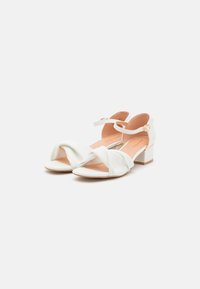 White heeled sandals with a twisted front design, ankle strap, and a block heel. Smooth texture with a peach-colored insole.