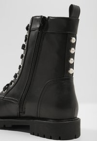 Černá kožená kotníková bota se zipem na boku, s šněrováním na přední straně, stříbrnými doplňky a perlovými akcenty podél podpatku pro zvýšení detailu.