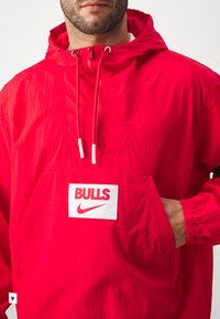Röd nylonpulloverjacka med mesh-textur, justerbar dragsko på huva, kenguruficka och djärv vit "BULLS" samt Nike-logga.