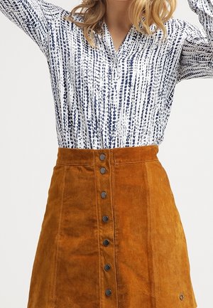 Woman wearing a white and navy patterned blouse tucked into a high-waisted brown suede button-front skirt.