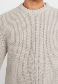 Beżowy sweter dzianinowy z prążkowanym okrągłym dekoltem, pokazany na osobie od ramion do połowy klatki piersiowej.