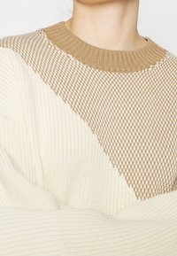 Osoba oblečená v krémově a hnědě pleteném svetru s texturovaným diamantovým vzorem na hrudi a žebrovanými manžetami a límcem.