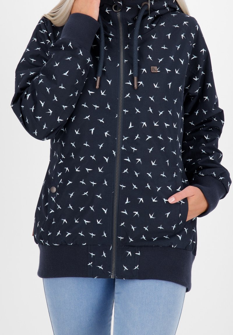 Kvinna i marinblå zip-up hoodie med vita fågelmönster, med ena handen i fickan och den andra handen nära ansiktet.