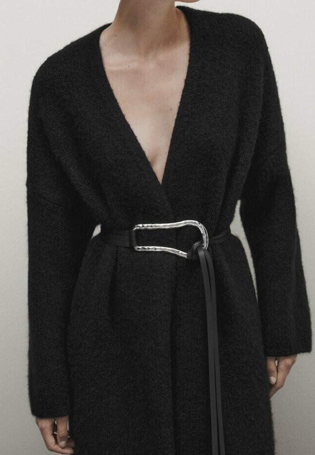 Femme portant un cardigan noir texturé avec des épaules tombantes, cintré à la taille par une ceinture noire avec une grande boucle argentée.
