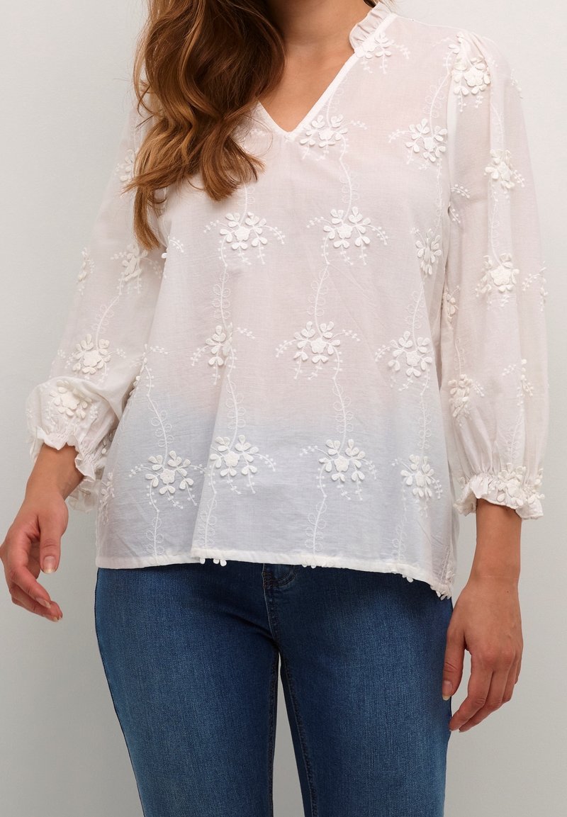 Femme portant un chemisier blanc à manches longues avec broderies florales et poignets volantés, associé à un jean bleu, sur un fond uni.