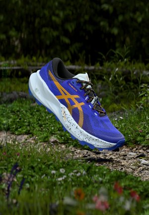 Paarse en witte hardloopschoen met oranje accenten, bovenwerk van gestructureerd mesh, gevoerde kraag en gespikkelde midsole. Zool met ontwerp gericht op grip.