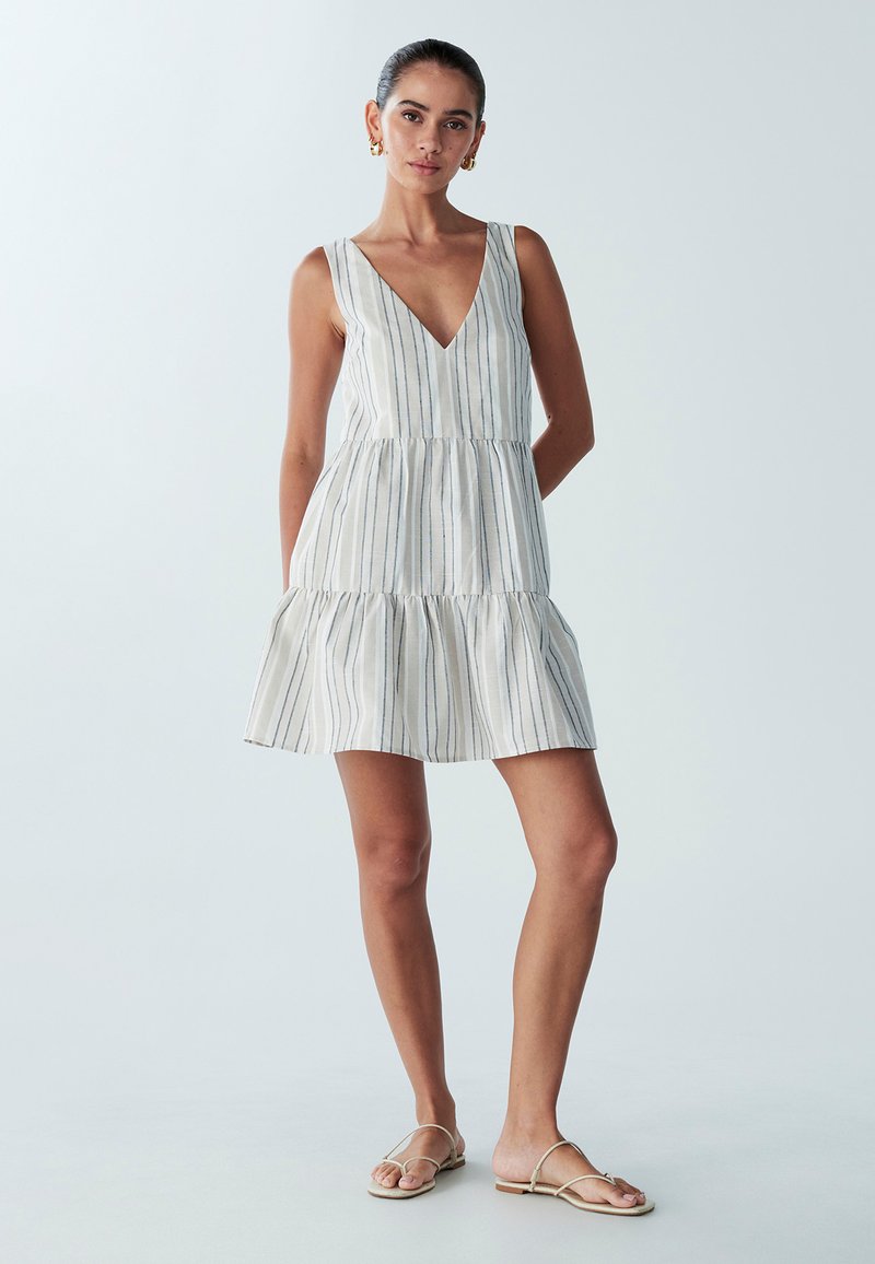 Paski mini sukienka z dekoltem w kształcie litery V, z warstwowym krojem, wykonana z lekkiego materiału z pionowymi niebieskimi i szarymi paskami. Dobrzona z beżowymi sandałami.