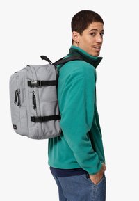 Zaino in tessuto grigio con spallacci, zip e fibbie nere. Presenta più scomparti e una forma rettangolare. Indossato su una spalla.