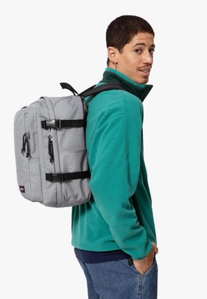 Mochila de tela gris con correas, cremalleras y hebillas negras. Presenta múltiples compartimentos y una forma rectangular. Lleva puesta en un hombro.