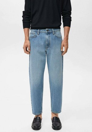 Model wears a black long-sleeve shirt and light blue denim jeans, paired with black loafers, against a neutral background.