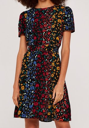 Mujer vestida con un vestido negro de manga corta y largo hasta la rodilla, con patrones florales multicolores en azul, rojo, amarillo y verde.