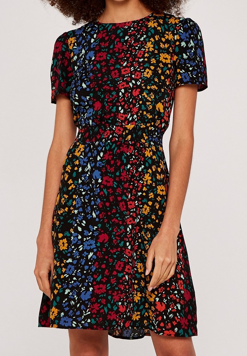 Femme portant une robe noire à manches courtes, arrivant au genou, avec des motifs floraux multicolores en bleu, rouge, jaune et vert.
