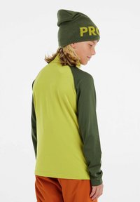 Lange mouw shirt in fel limoengroen met donkergroene mouwen, gemaakt van gladde stof. Gecombineerd met een olijfgroene beanie met gele tekst.