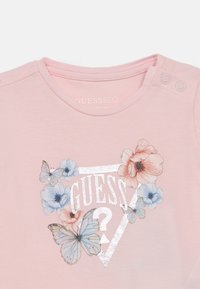 Ροζ βαμβακερό T-shirt με ασημί λογότυπο "GUESS", διακοσμημένο με μπλε και ροζ λουλουδάτα σχέδια και πεταλούδες. Κουμπιά στο ώμο για κούμπωμα.