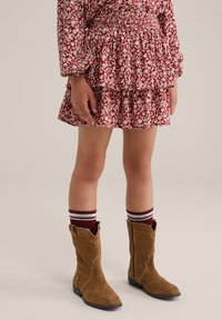 Klädd i en röd och vit blommönstrad klänning med volangsläpp, tillsammans med bruna mocka cowboy kängor och sockar med burgunderrand.