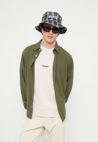 Jauns vīrietis, valkājot melnu rakstainu cepuri ar plata malka, melnas saulesbrilles, zaļu atvērtu kreklu pār bēša krāsas JJ Studios T-kreklu un bēša krāsas bikses uz balta fona.