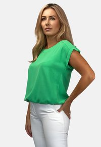 Zielona bluzka z krótkim rękawem wykonana z gładkiego materiału, o luźnym kroju. Projekt zawiera podwinięte rękawy i okrągły dekolt. Noszona z białymi spodniami.