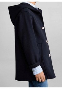 Námořnický kabát ze směsi vlny s kapucí, s stříbrnými knoflíky, bočními kapsami a volným střihem. Viditelná vrstvená světle modrá košile pod ním.