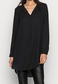 Svart långärmad blus med notchkrage, slät textur, knäppte ärmmuddar och en något förlängd bakre fåll. Lämplig för lagerpålägg.