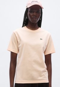 Ferskenfarvet bomuldst-shirt med korte ærmer, rund hals og et grønt Lacoste-logo på brystet. Glat tekstur med en afslappet pasform.
