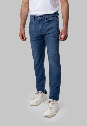 Uomo che indossa jeans blu slim fit con rinforzi sulle ginocchia e sneakers bianche, in piedi su uno sfondo grigio uniforme.