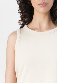 Top sin mangas beige con escote redondo, fabricado en una tela suave. Presenta un detalle de costura mínima a lo largo del escote y las sisas.