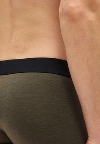 Κοντινό πλάνο της κάτω μέσης και του χεριού ενός ατόμου που φοράει υφασμάτινο λαδί παντελόνι με μαύρη μέση.