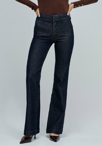 Jeans azules de cintura alta, con pierna acampanada, hechos de tela de mezclilla. Cuentan con un cierre de dos botones y pasadores para cinturón. Se combinan con tacones de punta.