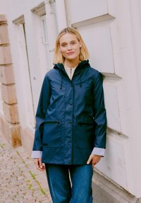 Veste imperméable marine avec capuche, fermeture à boutons sur le devant, deux poches zippées à l'avant et poignets rayés contrastants. Texture lisse, coupe décontractée.
