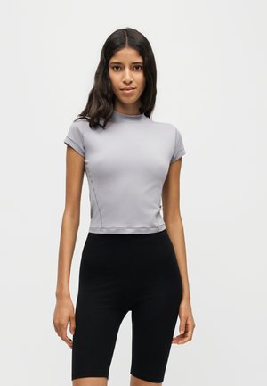 Ung kvinde med langt mørkt hår iført en lysegrå tætsiddende kortærmet top og sorte shorts til knæet, står foran en ensfarvet baggrund.