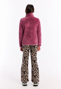 Žena okrenuta leđima, u bufnom ljubičastom kaputu, hlačama s uzorkom leopard, te crno-bijelim cipelama, na bijeloj pozadini.