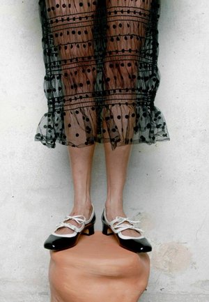 Piernas con falda negra transparente de lunares y zapatos de tacón bajo en blanco y negro, de pie sobre un pedestal redondeado de terracota frente a una pared clara.