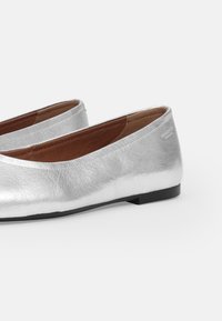 Silverfärgade balettflatser i texturerat metalliskt material, med en låg svart sula och brunt inre foder. Rund tådesign.