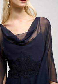 Mörkblå klänning med genomskinlig draperad halsringning och trumpetärmar, prydd med intrikata svarta spetsdetaljer vid midjan. Elegant och texturerad tyg.