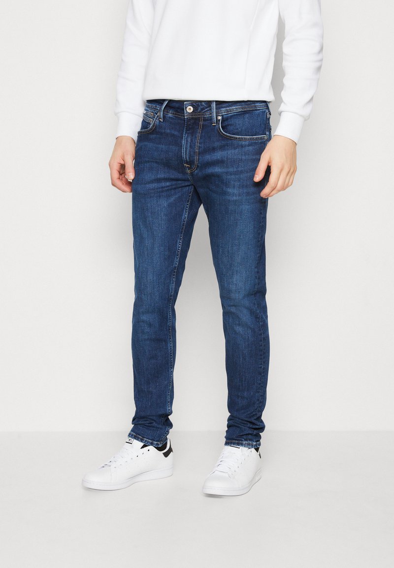 Mörkblå denimjeans med smal passform, fem fickor och subtila sömnadsdetaljer, tillsammans med vita sneakers.