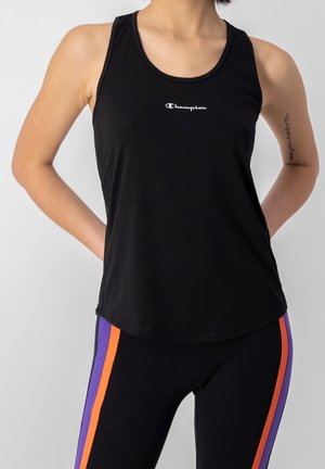 Person trägt schwarzen ärmellosen Champion-Top und schwarze Leggings mit lila und orangefarbenen Seitenstreifen, steht mit den Händen hinter dem Rücken.