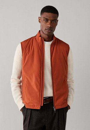 Mann trägt eine orange ärmellose Zip-Weste über einem weißen Strickpullover und dunklen Hosen, steht mit Händen in den Taschen vor einem schlichten Hintergrund.