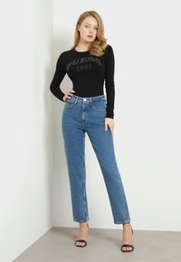 Čierny top s dlhým rukávom s grafikou "GUESS 1981" a modrými nohavicami s vysokým pásom. Model stojí proti svetlej stene, na nohách má čierne sandále na podpätku.