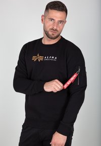 Sweatshirt noir en coton avec col rond, présentant un logo doré sur la poitrine et une poche à zip sur la manche avec une étiquette rouge.