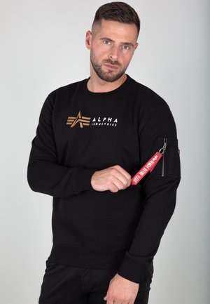 Sweatshirt noir en coton avec col rond, présentant un logo doré sur la poitrine et une poche à zip sur la manche avec une étiquette rouge.