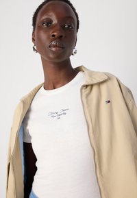 Giacca zip beige chiaro sopra una t-shirt bianca con testo ricamato blu. La t-shirt ha un collo rotondo. Orecchini a cerchio in argento visibili.