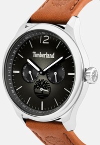 Timberland kell musta kelladialiga, hõbedase korpuse ja pruuni nahksanga ning päeva/kuupäeva aladialidega, mille all on logo.