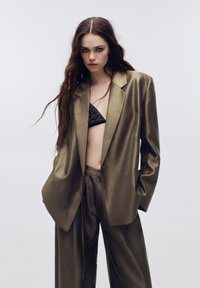 Costume en satin vert olive avec un blazer ample et un pantalon à jambes larges. Texture brillante remarquable, associé à un soutien-gorge noir brodé.