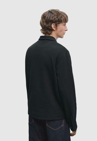Polo noir à manches longues avec col, texture côtelée et coupe légèrement décontractée. Présente des détails cousus au dos et aux poignets.