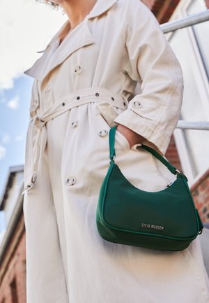 Borsa a tracolla in finta pelle verde con forma arrotondata, accessori in argento e logo impresso. Indossata con un trench coat color beige chiaro, che mette in evidenza entrambi gli oggetti.