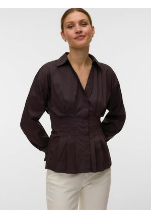 Frau trägt eine dunkelbraune, langärmlige, plissierte Bluse mit tiefem V-Ausschnitt und weiße Hose, steht mit den Händen hinter dem Rücken.